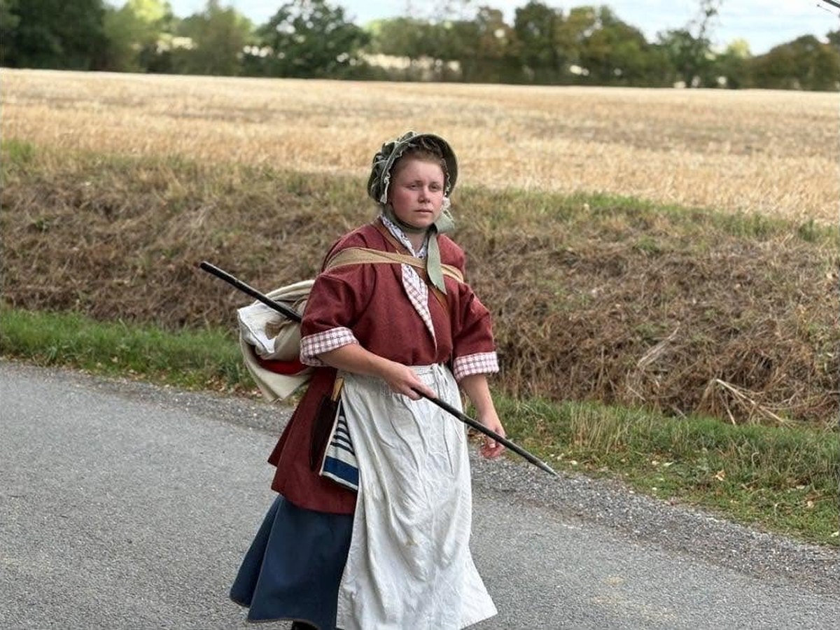 A woman in 1770s civilian dress walking with staff and pack