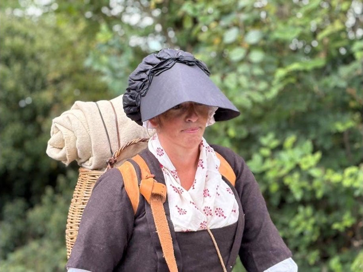 A woman in 1770s dress carrying a wicker basket on her back