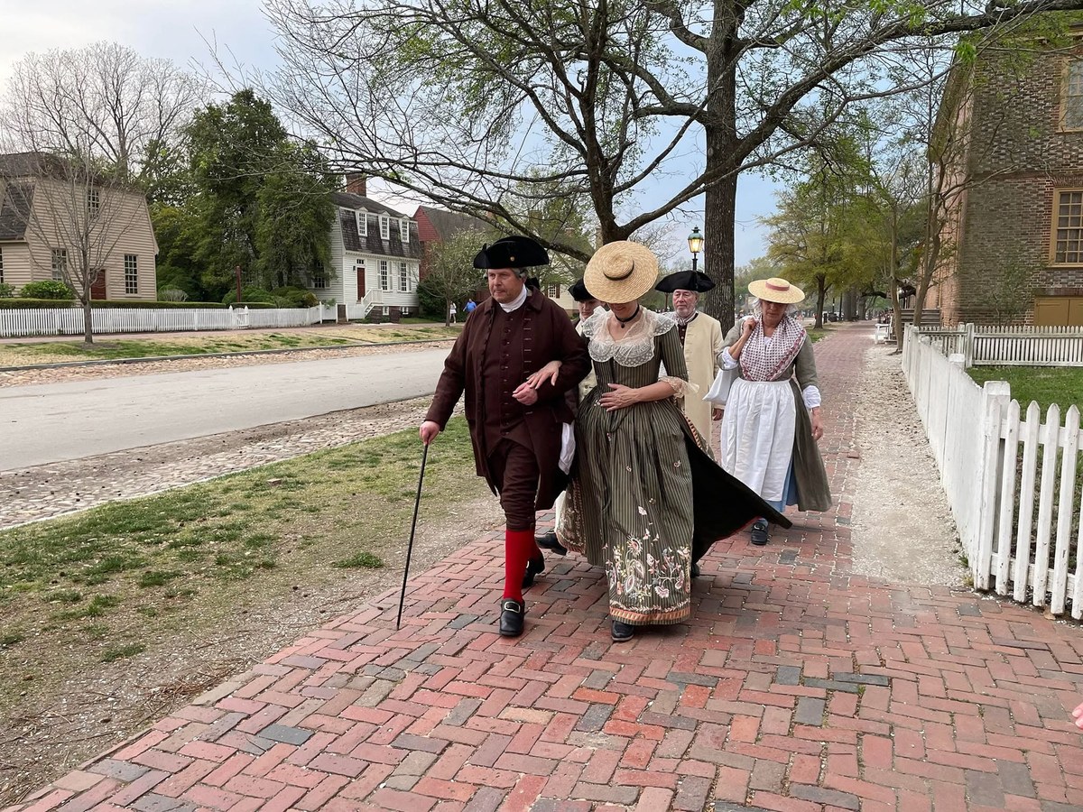 A gentleman and ladies in 1770s dress walking along a brick path